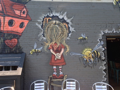 [A larger than life little girl standing on a step ladder, back to the viewer, looking into a hole, with some bees buzzing around her] If I didn't know better, these “honey bees” look more like “killer hornets.”