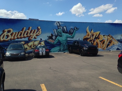 [A mural, convering the entire side of a building, with the word “Buddy's” on the left, the word “Detroit” on the right, and in the middle, Atlas, with the city skyline of Detroit on his back, contemplating a pan of Buddy's Pizza held in his right arm] ´It's a deep-dish square pizza with the sauce on top, and it's insanely delicious!